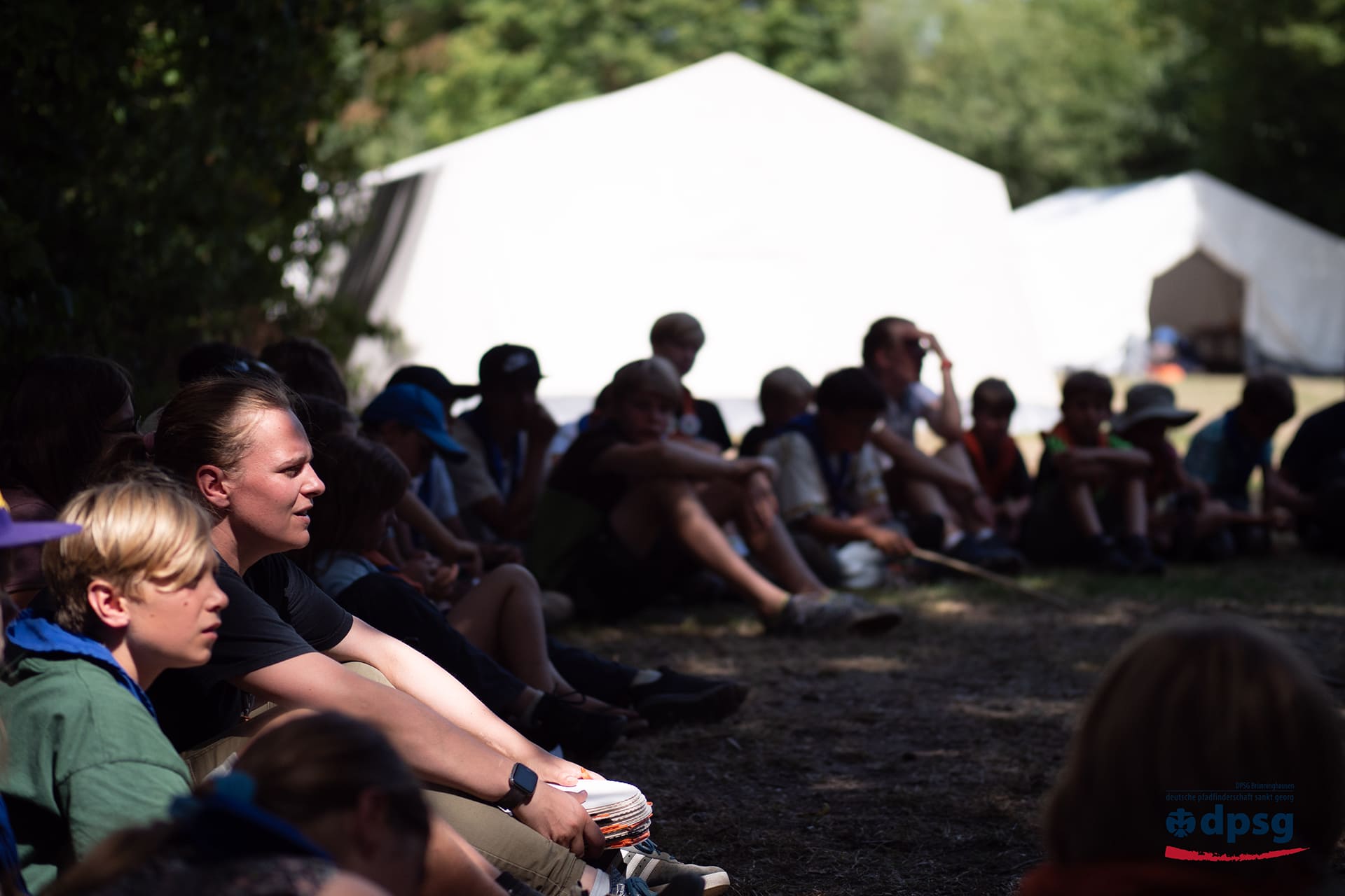Kinder, Jugendliche und Leitende sitzen im Zeltlager in einer Runde vor weißen Zelten